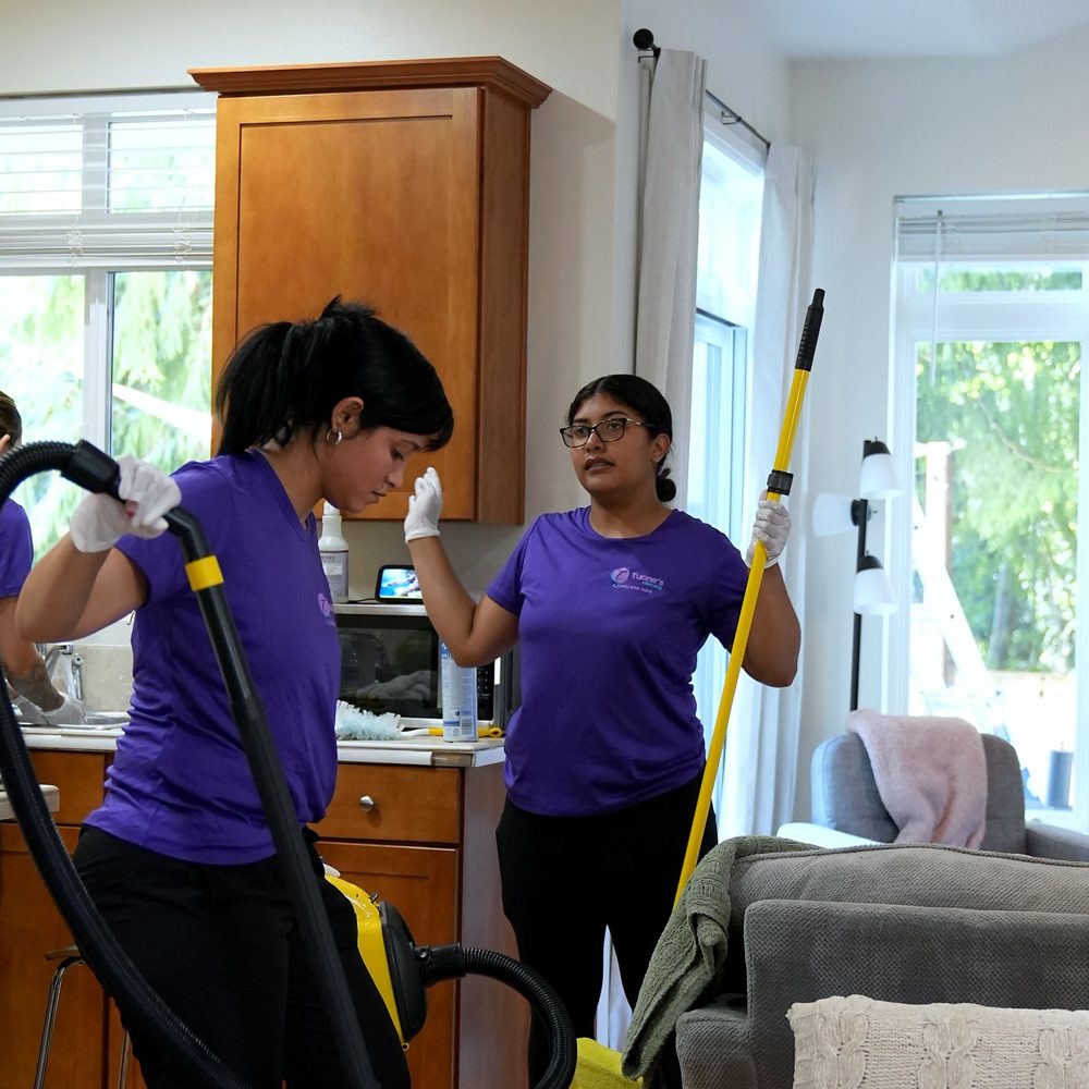 Two team members cleaning a kitchen and living room, wearing gloves and shoe covers